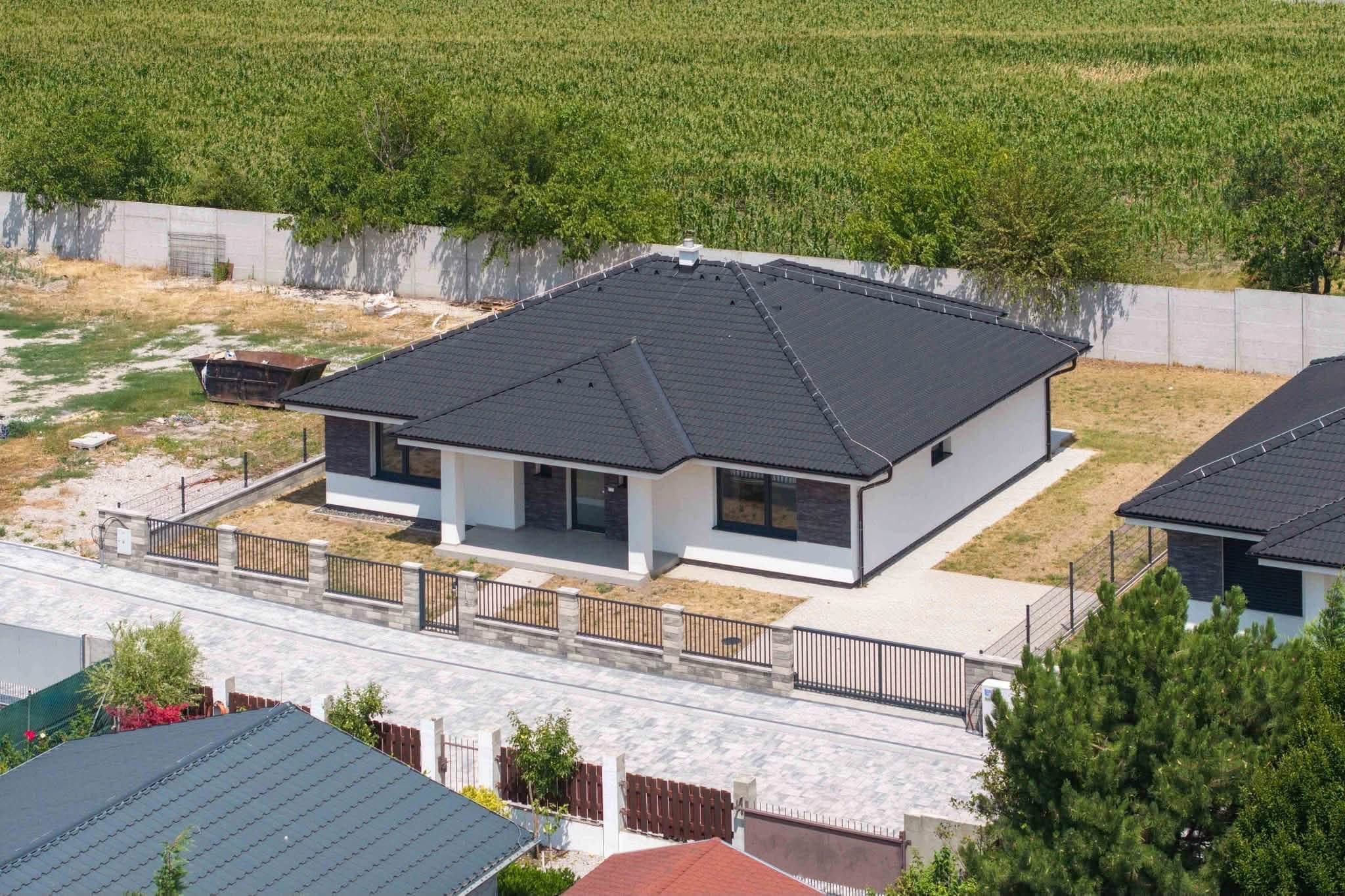 Sötét kontyolt tetős, fehér homlokzatú bungaló, drónfelvétel az utca felől
